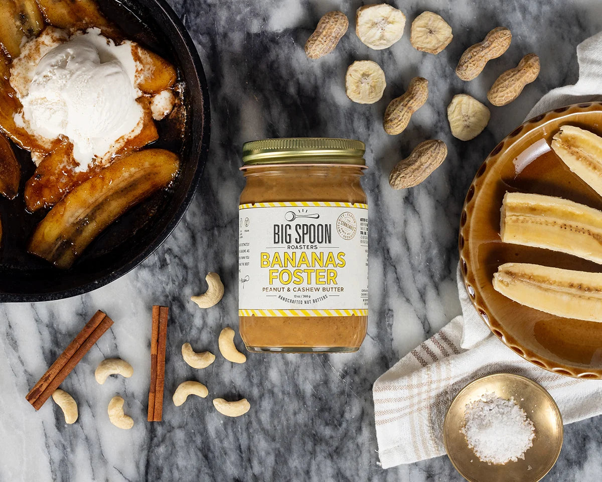 A jar of Big Spoon Bananas Foster peanut butter and cashew butter sits on a marble surface surrounded by banana slices, peanuts, cinnamon sticks, cashews, and dessert dishes.