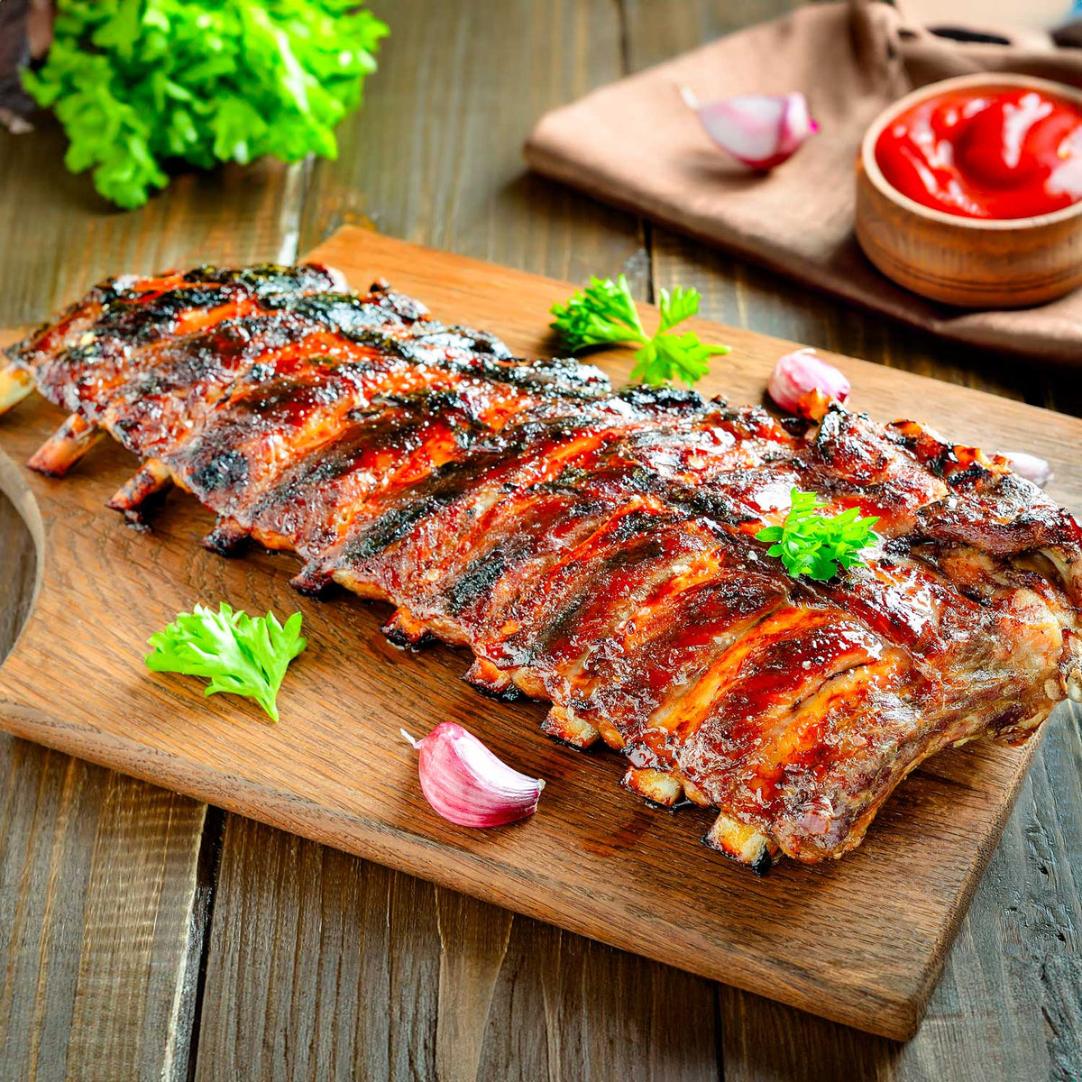 Grilled ribs on a wooden cutting board with garnishes on a rustic wooden table.