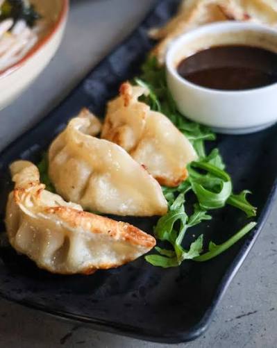 Pan-fried dumplings with crispy golden-brown bottoms served on a black rectangular plate with green arugula and a small white dipping sauce bowl.