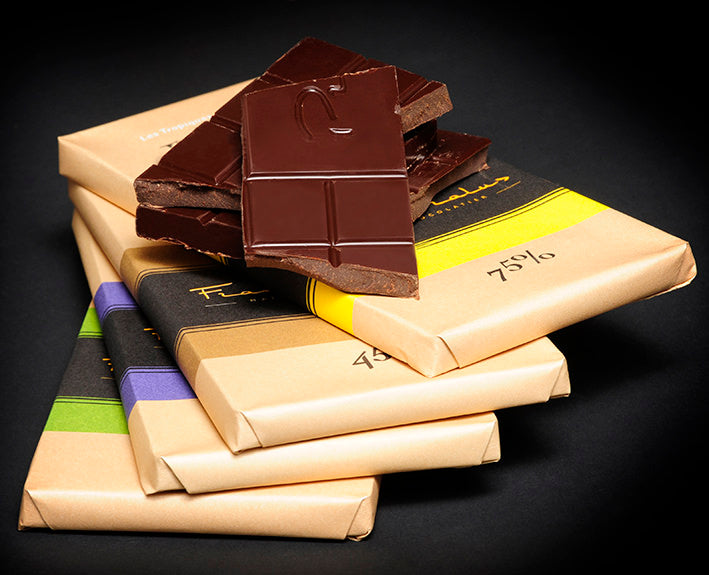 A stack of wrapped chocolate bars with one broken piece on top, photographed on a dark studio background. The wrappers are kraft paper with black bands and colored accents in yellow, purple, and green, and the top piece shows dark chocolate with a logo embossed