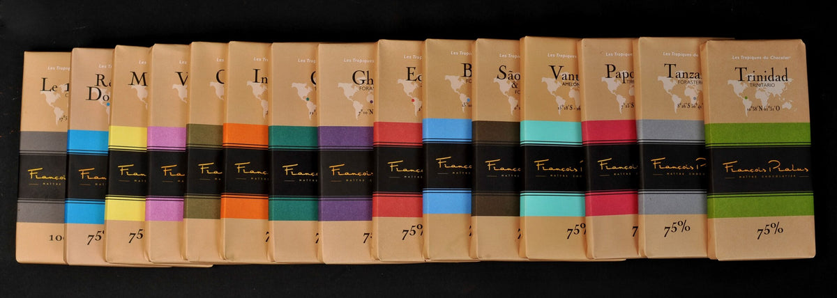 Row of François Pralus single-origin 75% dark chocolate bars standing upright on a dark surface, showing origin labels including Madagascar, Venezuela, Ghana, Ecuador, Brazil, São Tomé, Vanuatu, Papua, Tanzania, and Trinidad