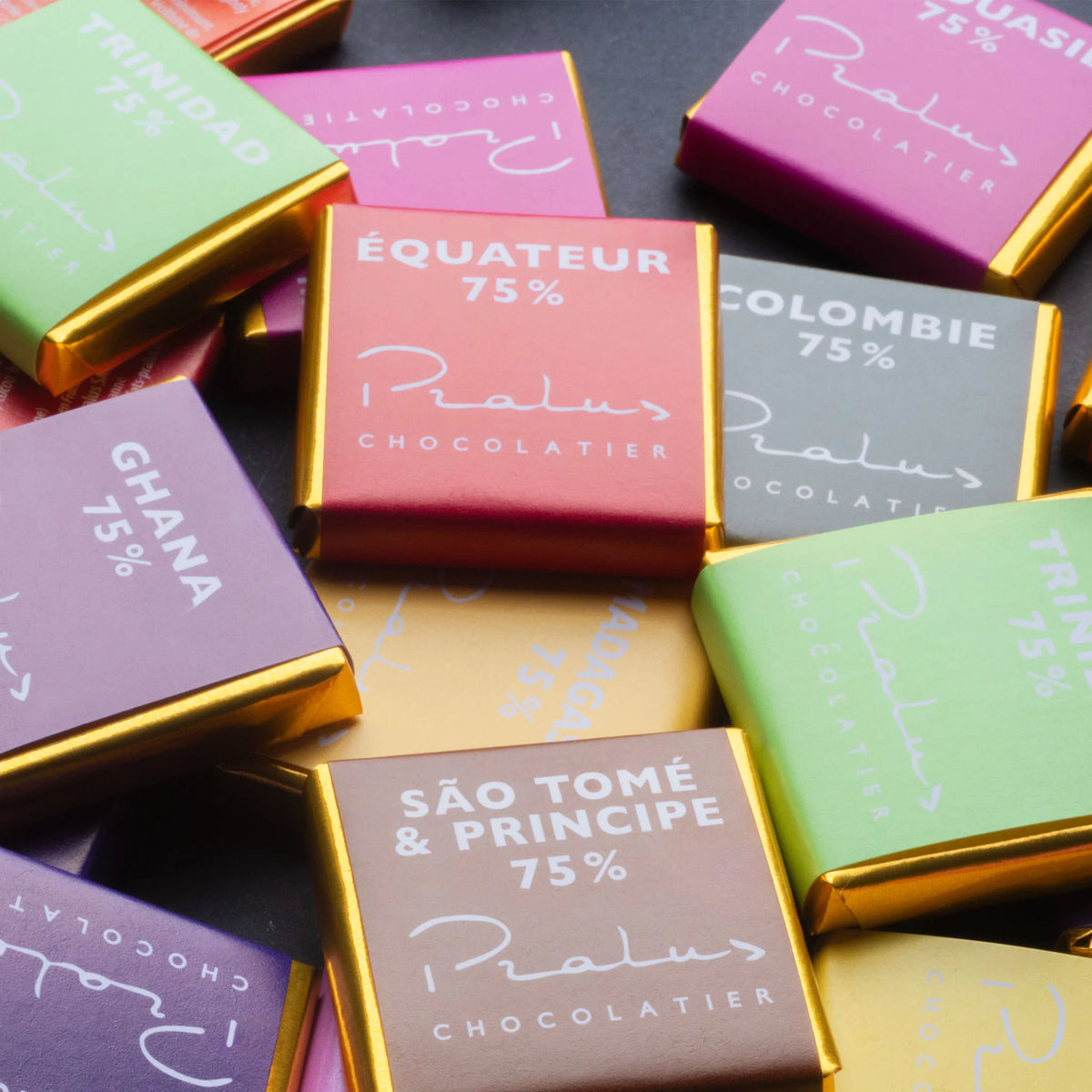 Overhead flat-lay of François Pralus mini single-origin 75% dark chocolate bars scattered on a dark surface — colorful wrappers labeled Équateur, Colombie, Ghana, São Tomé & Principe, and Papouasie with Pralus Chocolatier branding