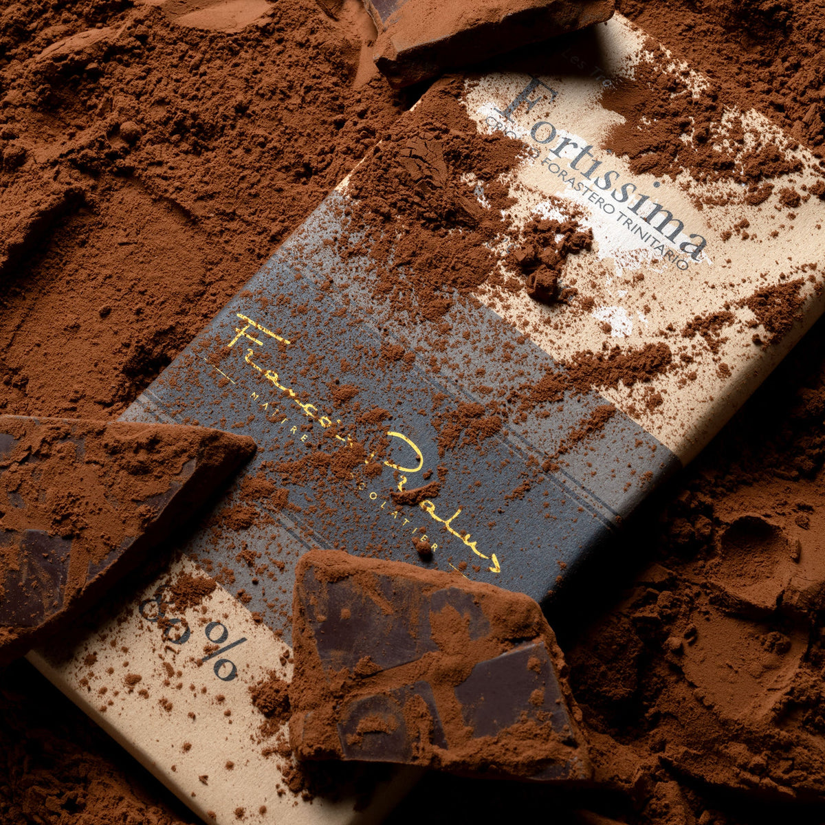 François Pralus Fortissima 80% dark chocolate bar in cream and purple packaging lying on a bed of cocoa powder with broken dark chocolate pieces scattered around it