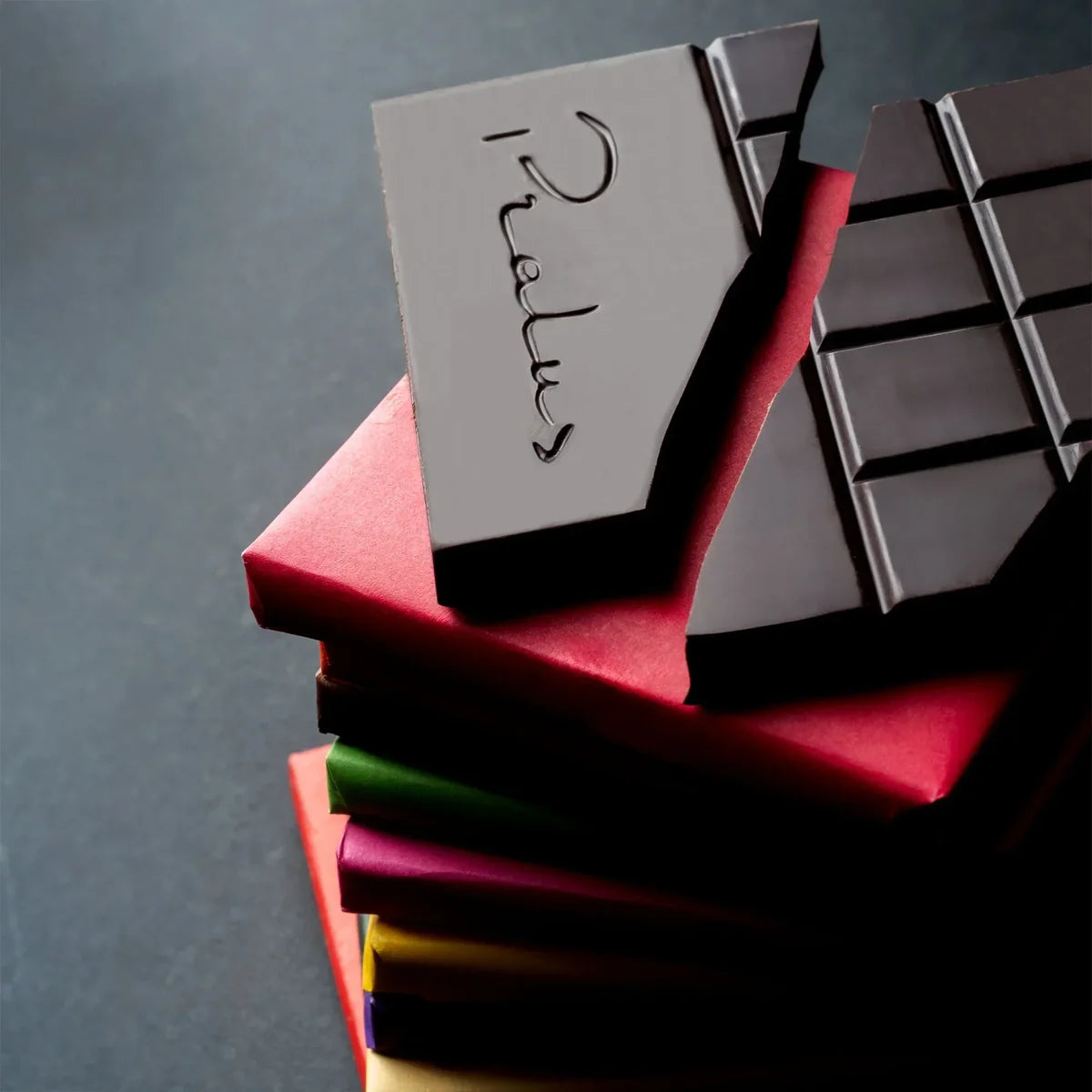 Stack of colorful François Pralus single-origin chocolate bar wrappers in red, green, and gold with a large dark chocolate bar leaning against them showing the embossed Pralus Chocolatier logo
