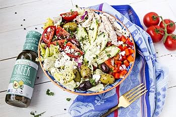 A bowl of fresh Greek salad with cucumber, tomatoes, olives, feta cheese, and grilled chicken, served with a bottle of Epiroikos Extra Virgin Olive Oil and a fork on a blue patterned cloth over a white wooden table.