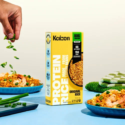 A box of Kaizen Protein Rice Original Rice stands on a blue surface with bowls of prepared rice and a hand sprinkling green onions into the scene. The product box is bright yellow with black and green text, and the surrounding food and kitchen props create a styled lifestyle setup.