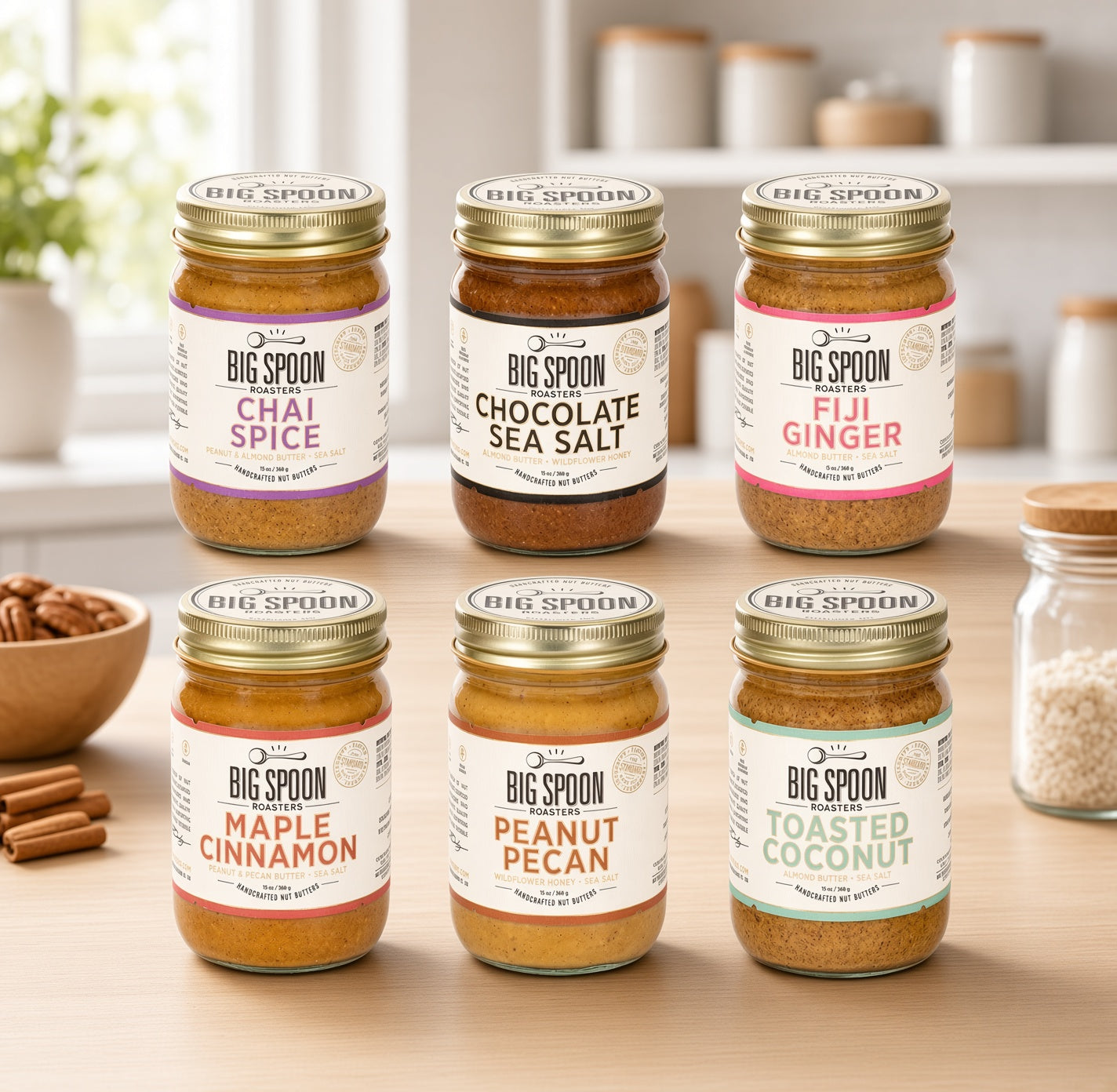 Six jars of Big Spoon Spice blends arranged on a wooden countertop in a bright kitchen setting. The jars include Chai Spice, Chocolate Sea Salt, Fuji Ginger, Maple cinnamon, Peanut Pecan, and Roasted Coconut.