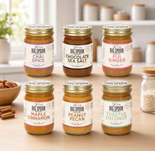 Six jars of Big Spoon Spice blends arranged on a wooden countertop in a bright kitchen setting. The jars include Chai Spice, Chocolate Sea Salt, Fuji Ginger, Maple cinnamon, Peanut Pecan, and Roasted Coconut.