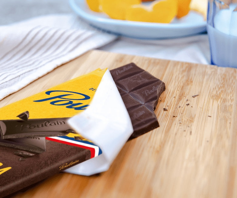 Poulain Noir Extra bar partially unwrapped on a wooden board with orange slices and milk in background
