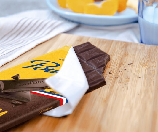Poulain Noir Extra bar partially unwrapped on a wooden board with orange slices and milk in background
