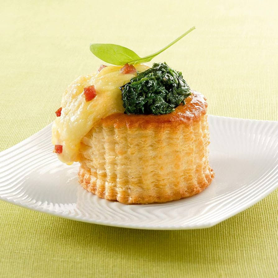A small baked pastry or cupcake on a white plate, topped with melted yellow cheese, a dollop of dark green spinach, and a small green leaf garnish, set on a light yellow-green textured background.