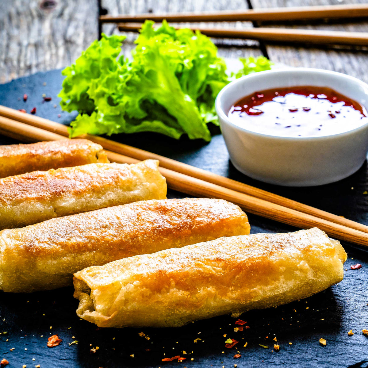 Spring rolls on a dark surface with lettuce, dipping sauce, and chopsticks.