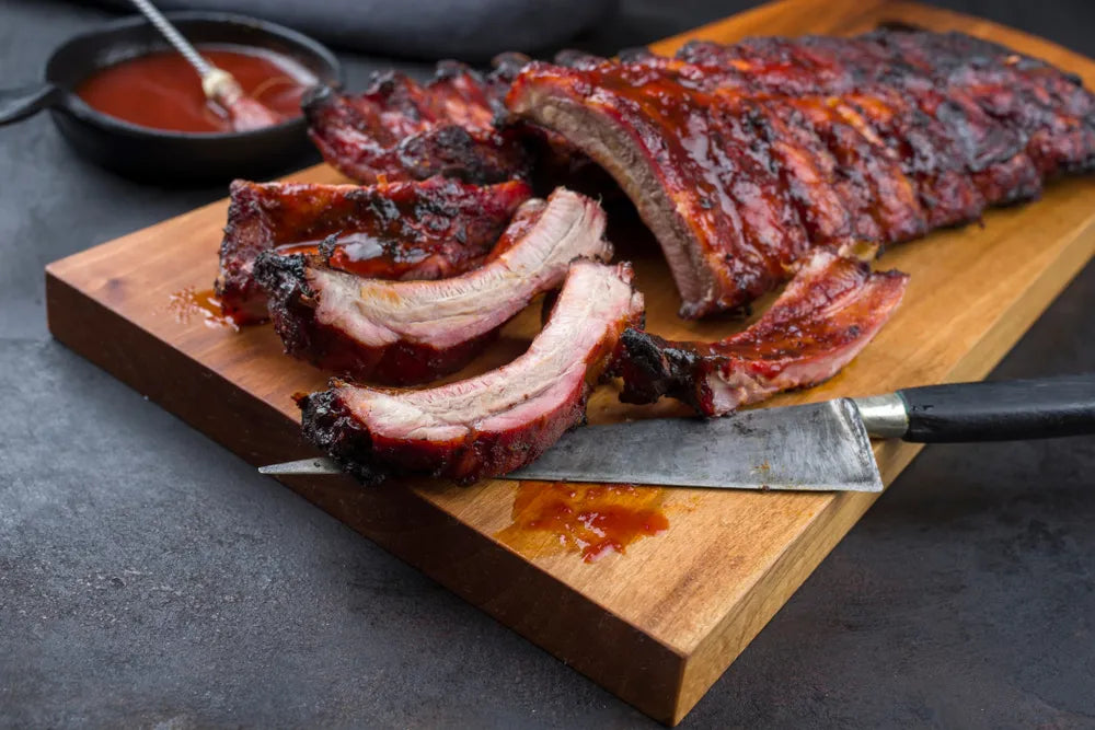 Sliced smoked ribs with a glossy red-brown barbecue sauce on a wooden cutting board, with a knife and a small bowl of sauce in the background.