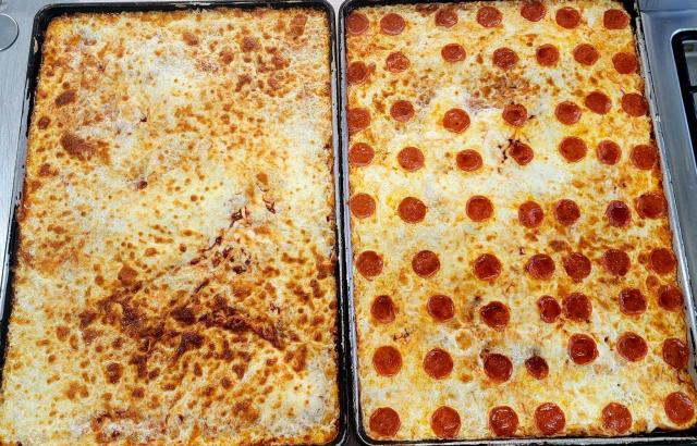 Two large rectangular baking pans filled with baked pizza on a metal surface. The left pan has a melted cheese topping with browned spots, and the right pan has a pepperoni pizza topping with many round slices visible across the surface.