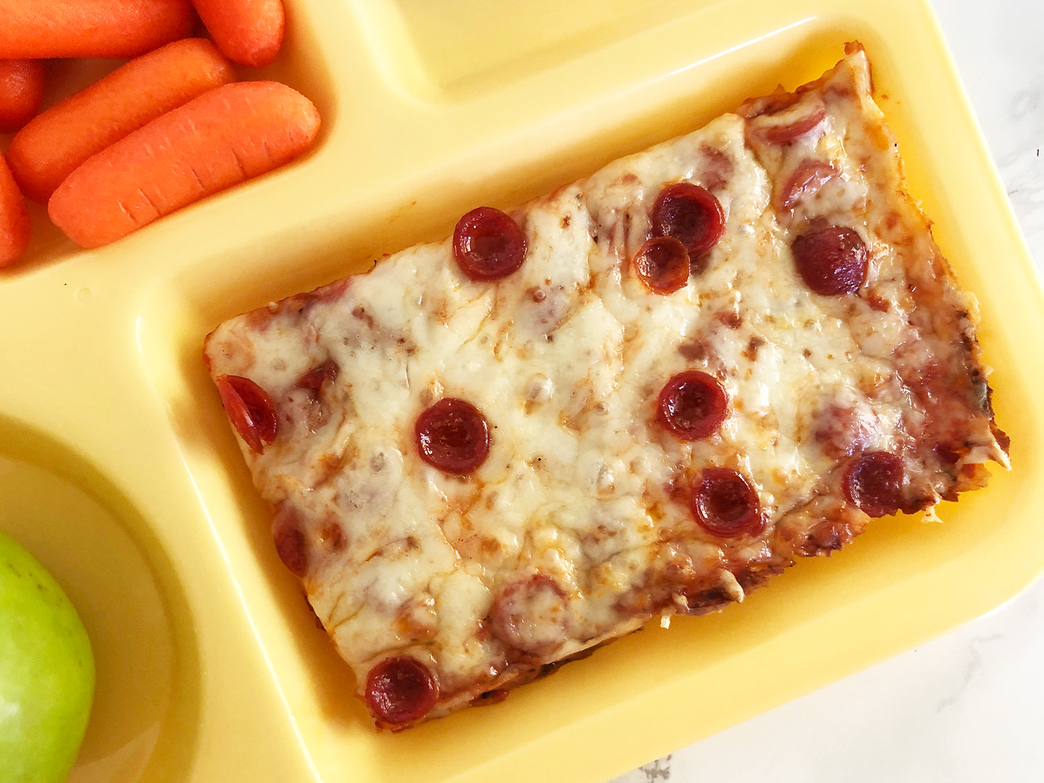 A close-up of a rectangular tray of cheesy pizza with pepperoni slices, served with baby carrots and a green apple in a yellow compartment tray.