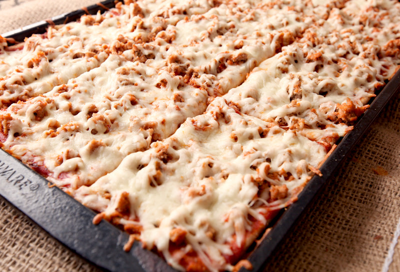 A close-up of a baked rectangular pizza or pasta dish in a dark baking pan, topped with melted white cheese and browned meat pieces, resting on a burlap surface.