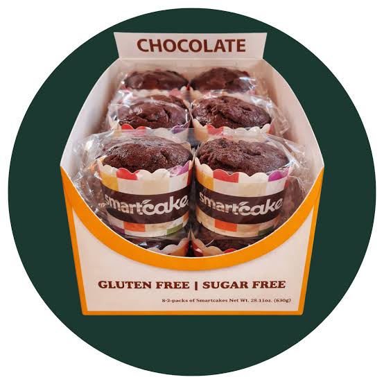 A box of six chocolate gluten-free sugar-free smartcakes in clear plastic wrappers, displayed inside an open white and orange cardboard box labeled 'CHOCOLATE' and 'GLUTEN FREE | SUGAR FREE'. The box is centered on a dark green circular background.