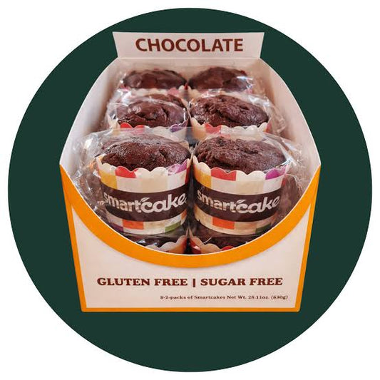 A box of six chocolate gluten-free sugar-free smartcakes in clear plastic wrappers, displayed inside an open white and orange cardboard box labeled 'CHOCOLATE' and 'GLUTEN FREE | SUGAR FREE'. The box is centered on a dark green circular background.