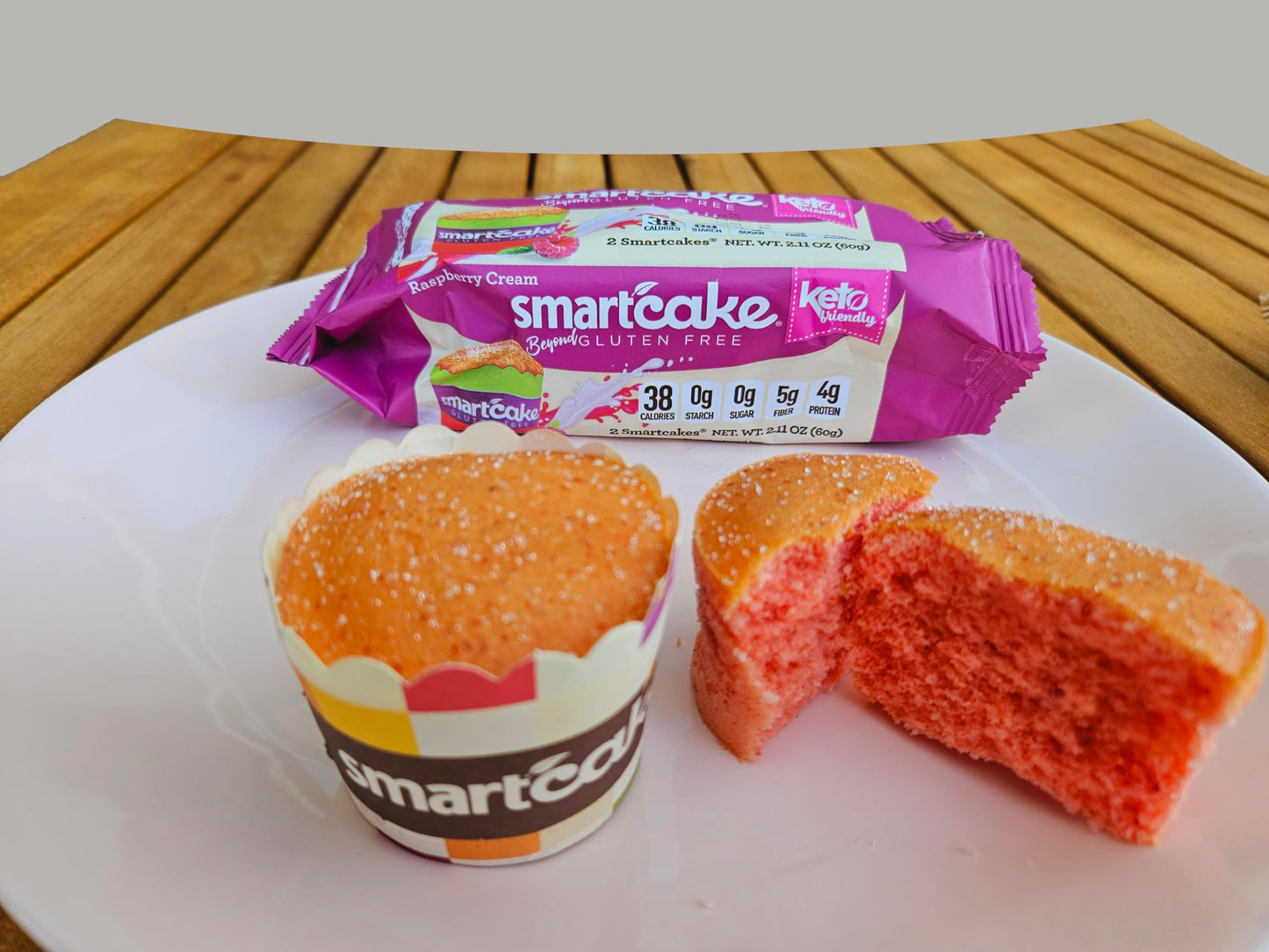 A smartcake super nutri-flourless muffin in a paper liner next to a broken piece showing the pink-orange crumb, with its purple and white wrapper behind it on a white plate over a wooden table.