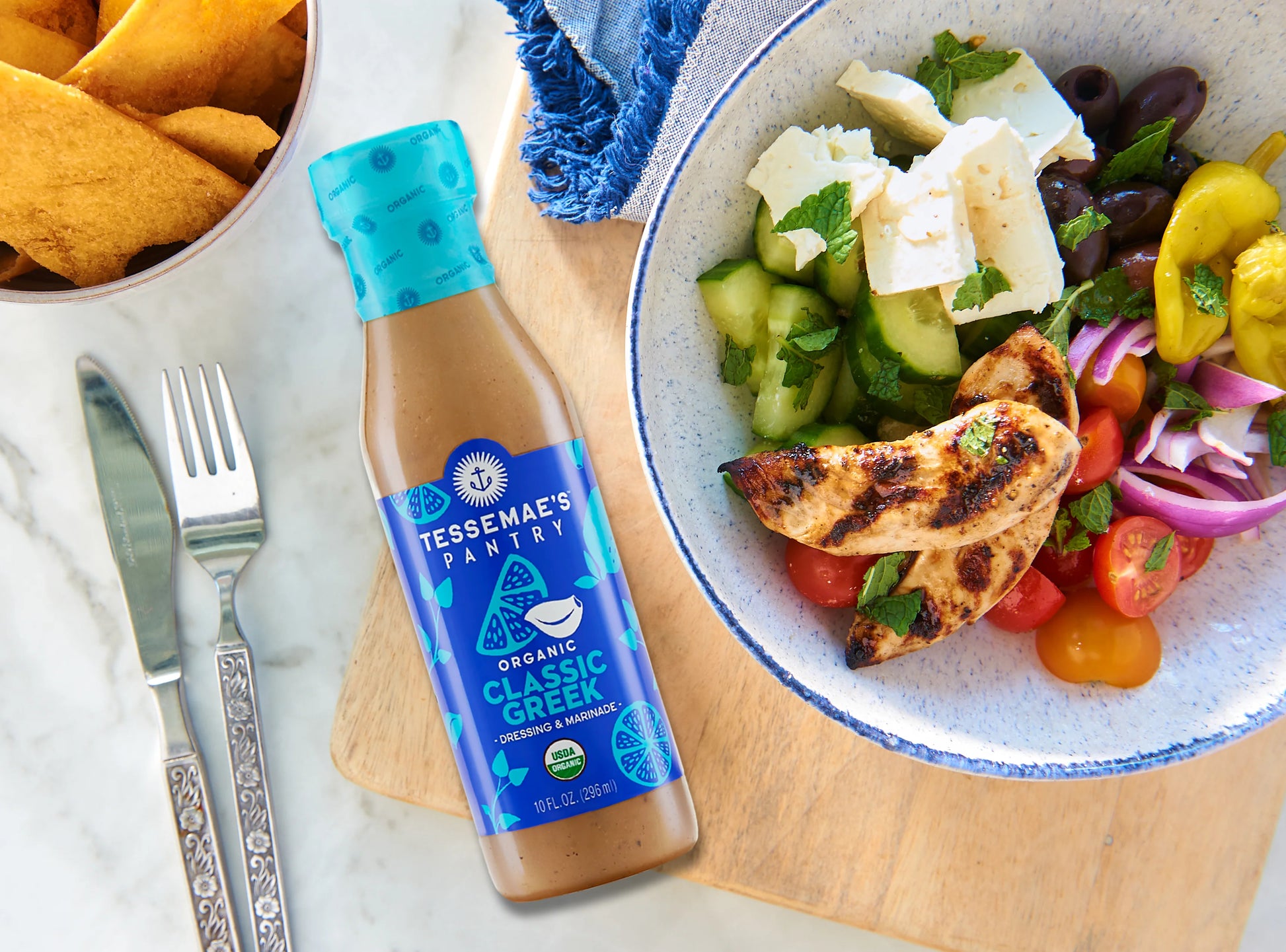 A bottle of Tessenae's Pantry Classic Greek dressing placed on a wooden board next to a bowl of salad with grilled chicken, tomatoes, cucumbers, feta cheese, olives, and a plate of chips on a white marble surface.