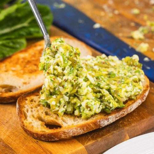 A close-up of a spoonful of green herb spread being spread onto a slice of toasted bread on a wooden cutting board, with another slice and a blue-handled knife in the background.