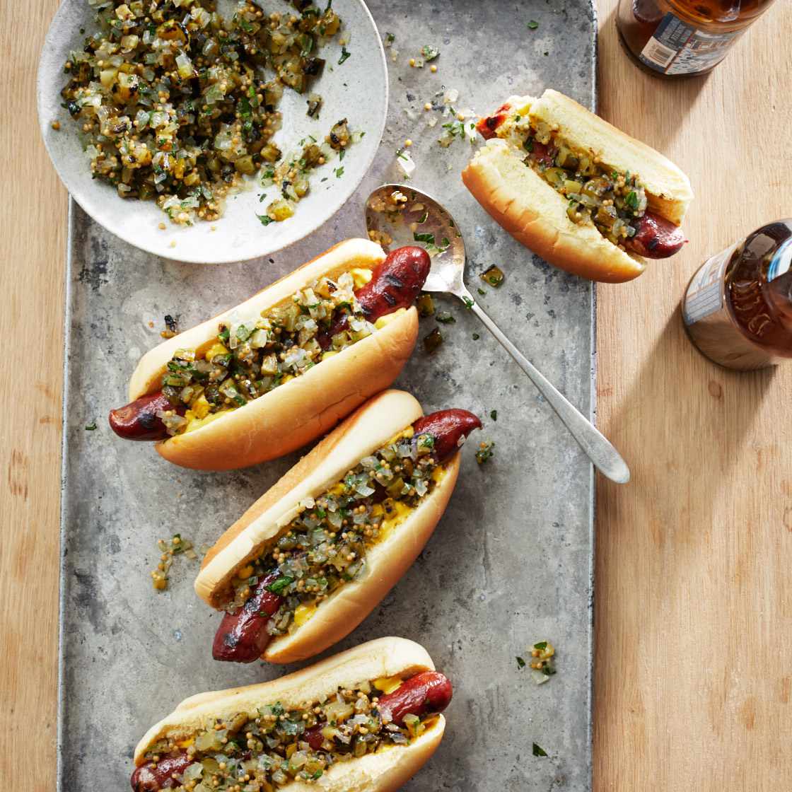 Four hot dogs topped with a chunky green and yellow relish, arranged on a gray metal serving board with a small plate of additional topping, a spoon, and two sauce bottles on a wooden table.