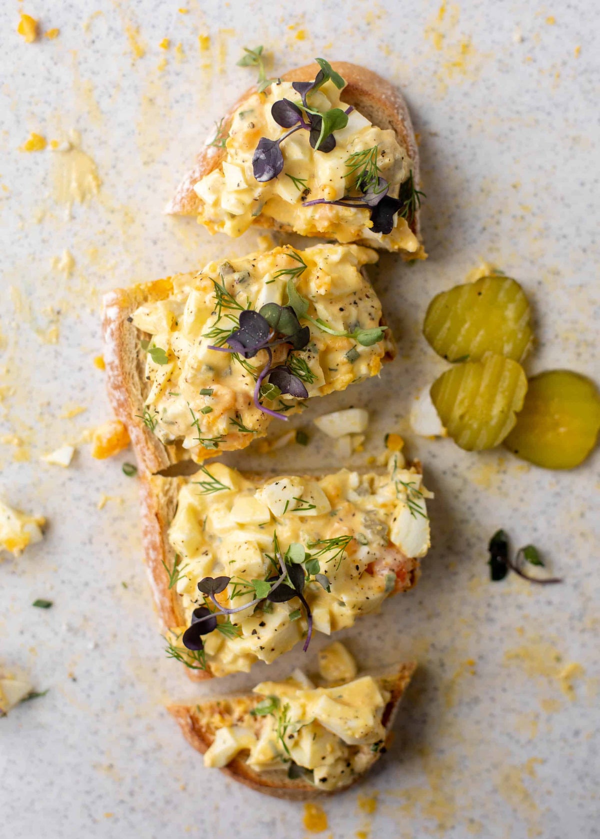 Four slices of toasted bread topped with a creamy yellow egg salad, garnished with microgreens, dill, and black pepper, arranged on a light speckled surface with two pickles on the right.